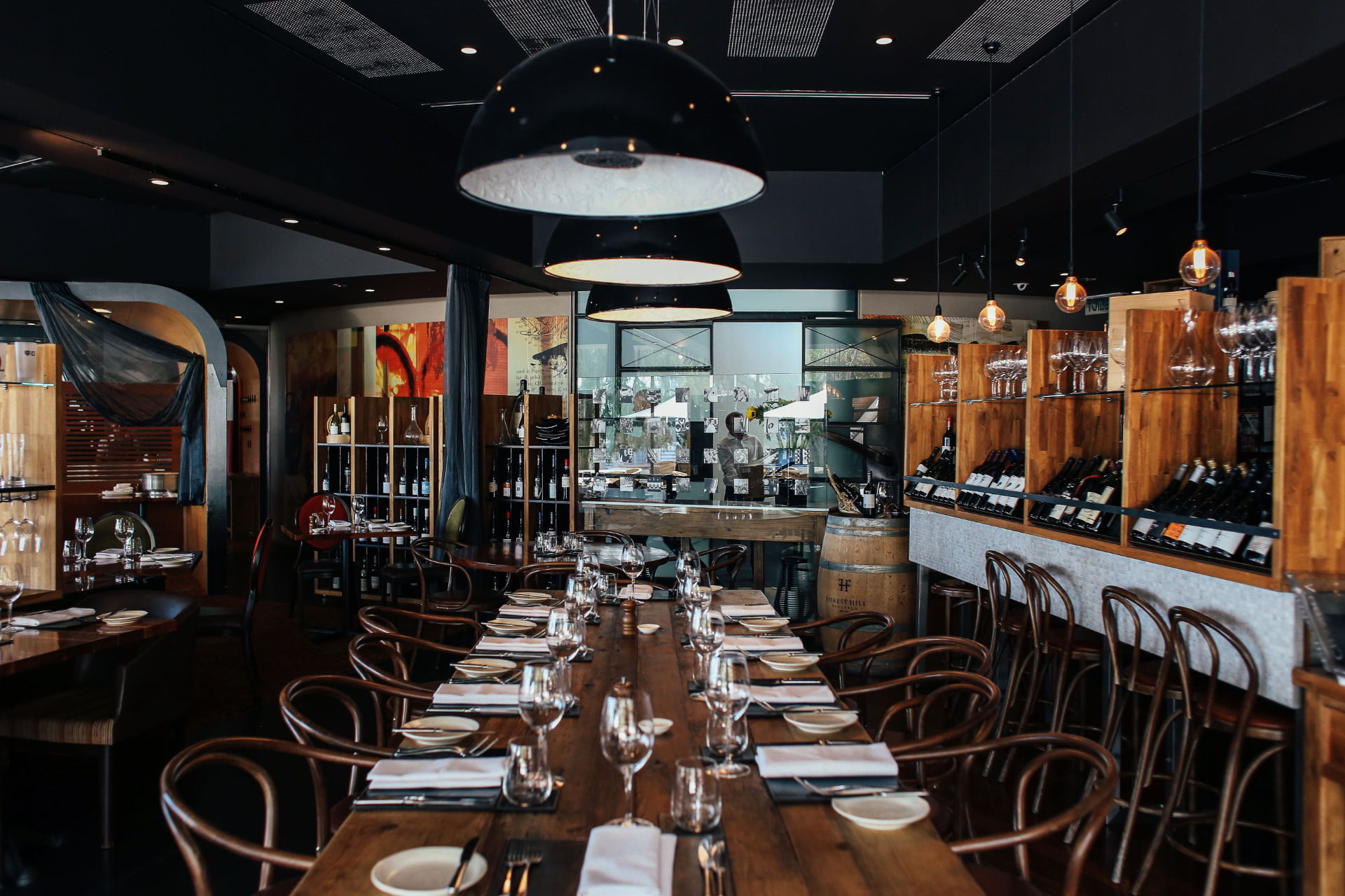 Moody restaurant interior with a long communal timber table, bentwood chairs and a wine wall